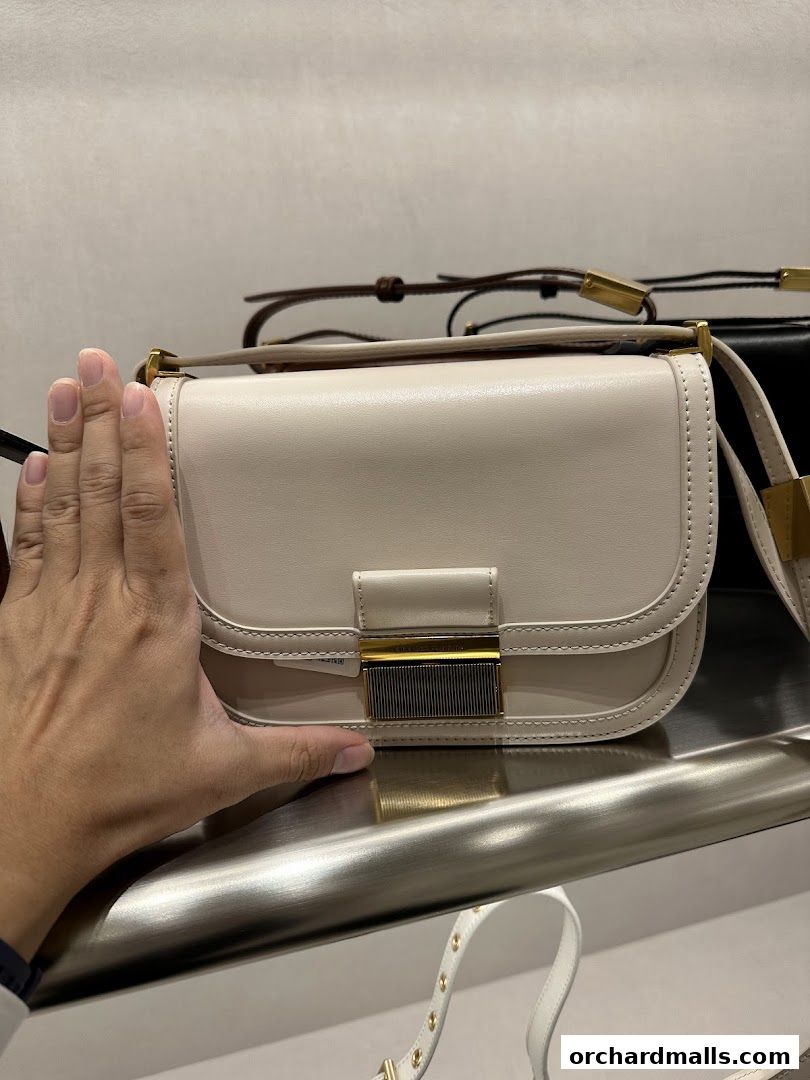 A hand is shown next to a cream-colored handbag with gold hardware.