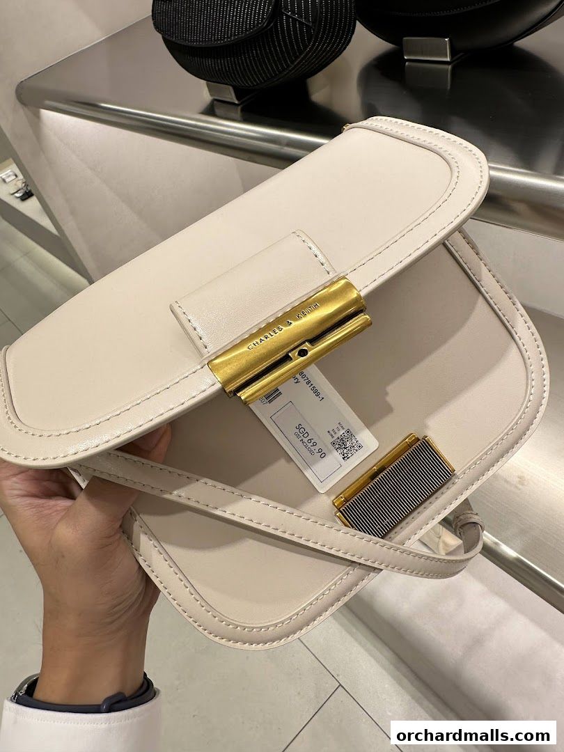 A hand holds a cream-colored Charles & Keith handbag with gold hardware.