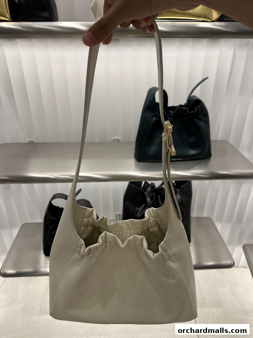 A hand holds a cream-colored ruched handbag in front of other handbags on display.