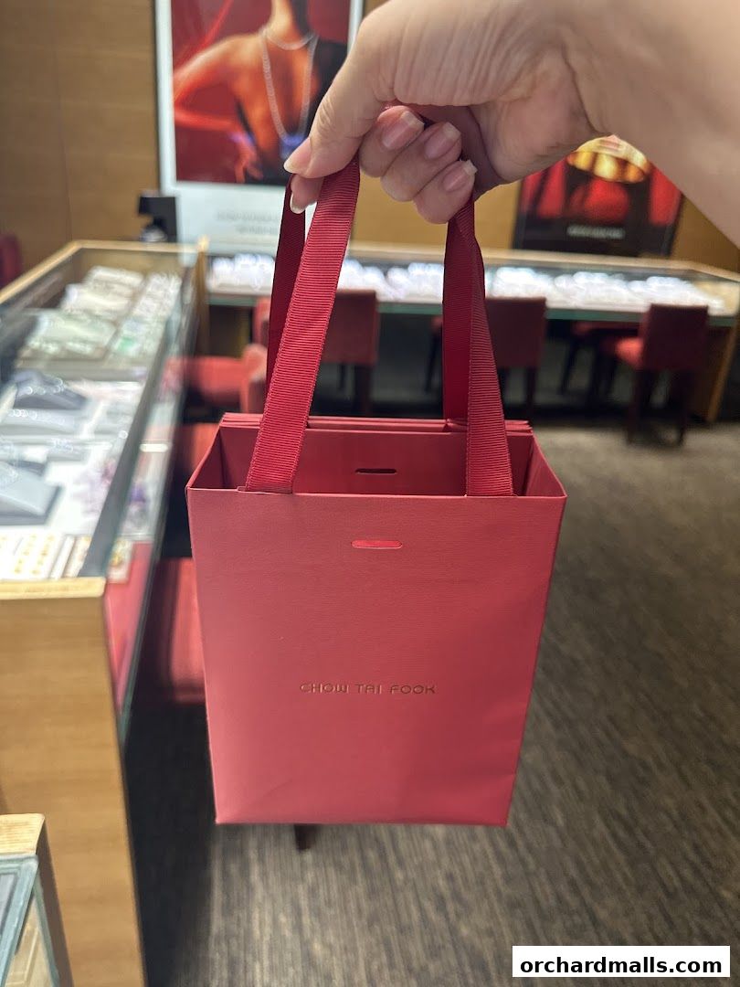 A hand holds a red Chow Tai Fook shopping bag inside a jewelry store.