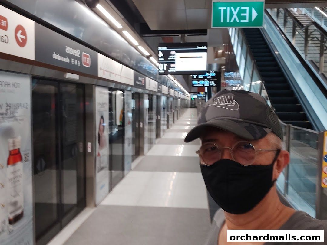 A person wearing a mask and cap in a subway station with trains and escalators.