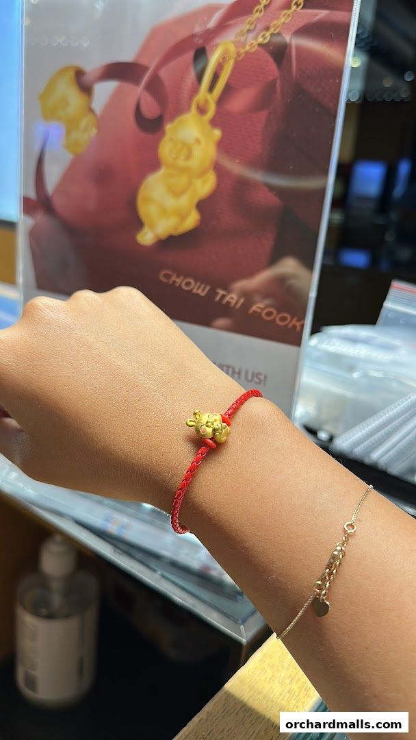 A hand wearing a red string bracelet with a gold pig charm, with a Chow Tai Fook display in the background.