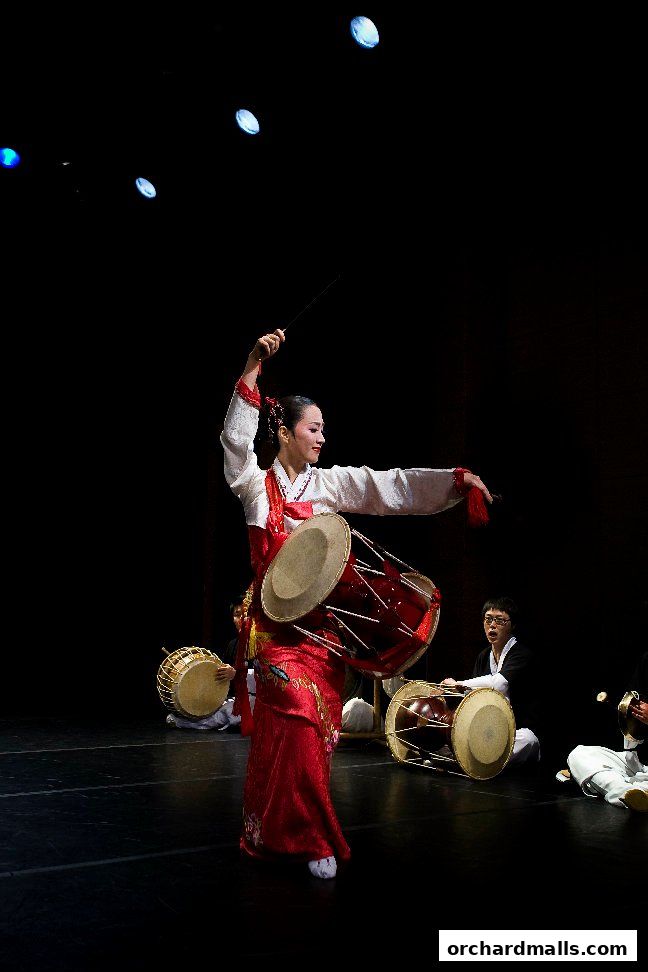 A woman in a red and white traditional Korean outfit dances with a drum.