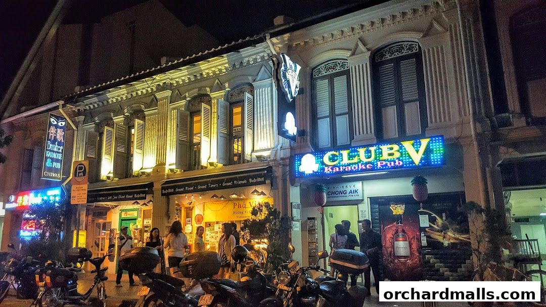 Night view of CLUB-V Karaoke Pub and other shops with neon signs and parked motorcycles.