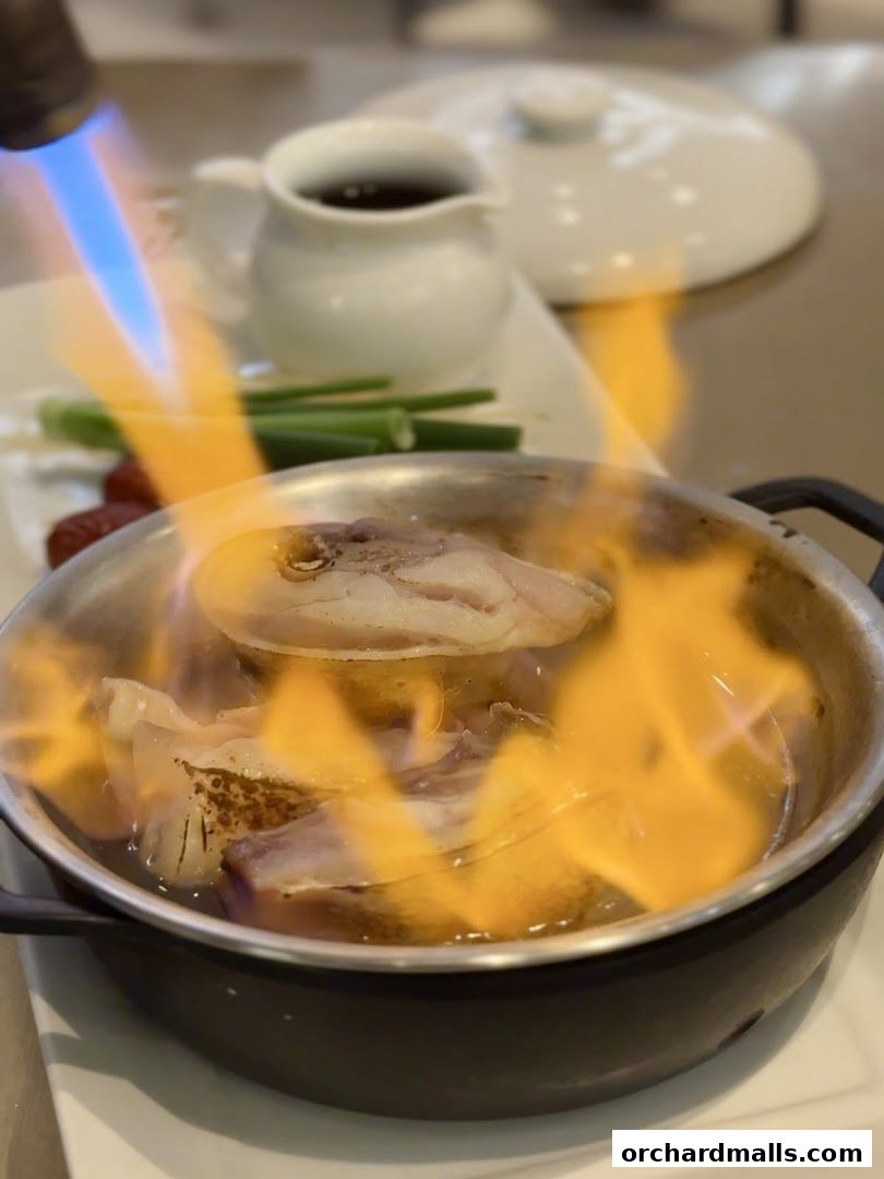 Fish being flambéed in a pot with a blowtorch, served with vegetables and sauce.