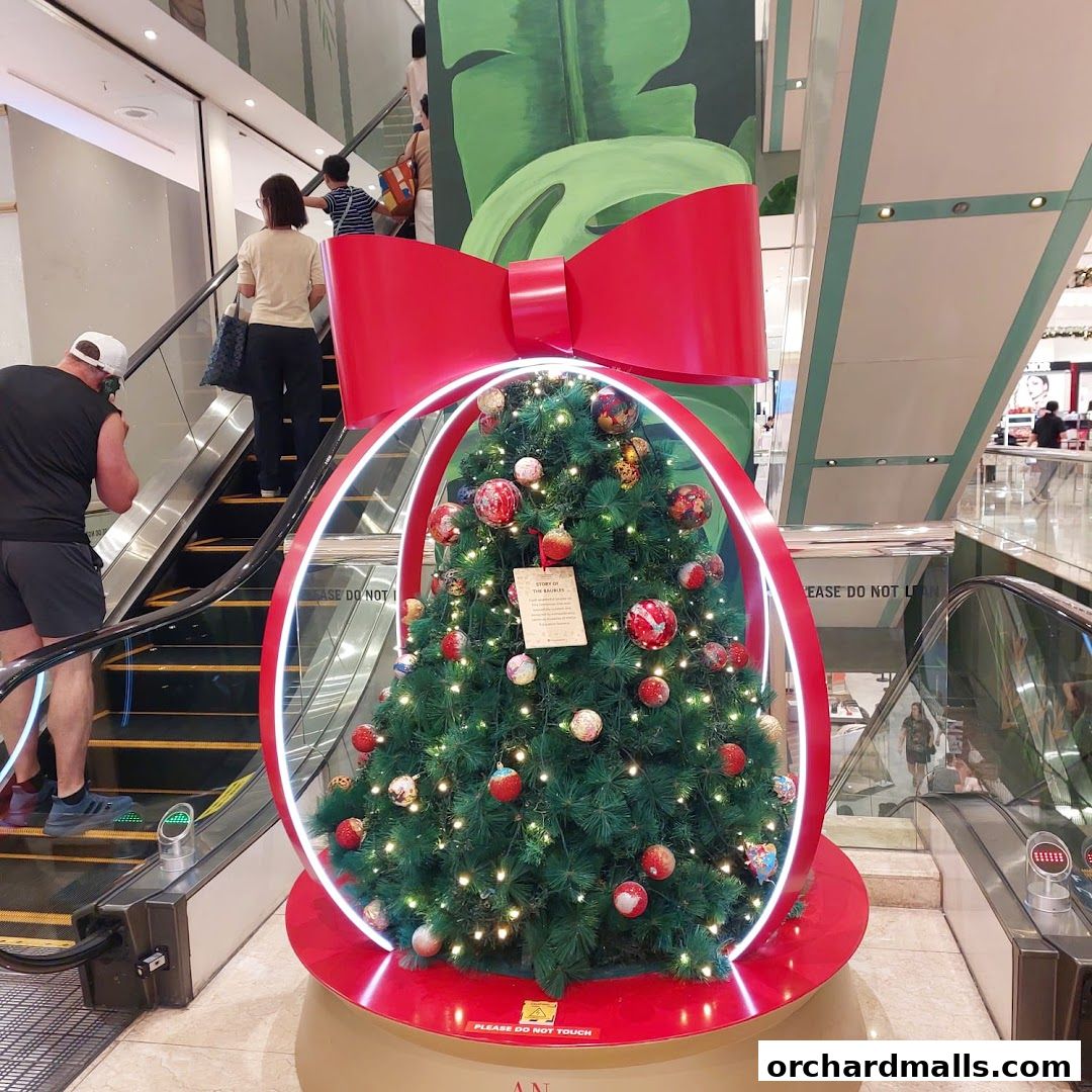 A decorated Christmas tree with a large red bow and lights, displayed near escalators in a mall.
