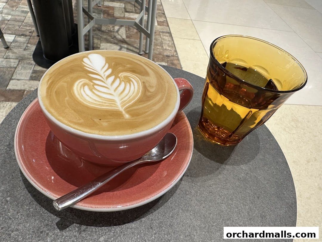 A beautifully crafted latte with intricate latte art sits on a pink saucer next to a glass of water.