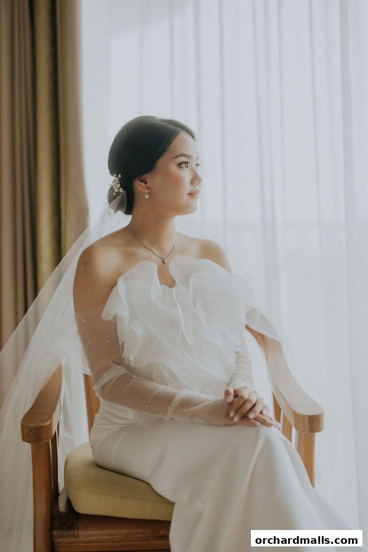 A bride in a white gown with a veil sits elegantly, looking out a window.