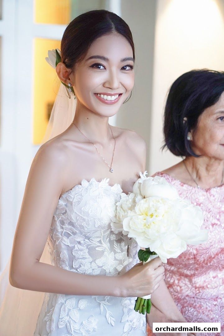 A smiling bride in a white gown holds a bouquet, with an older woman beside her.
