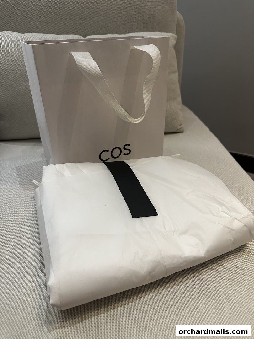 A white COS shopping bag and a garment bag with a black stripe on a sofa.