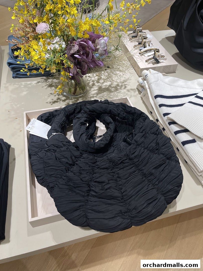 A black ruched tote bag is displayed with a bouquet of flowers and striped sweaters.