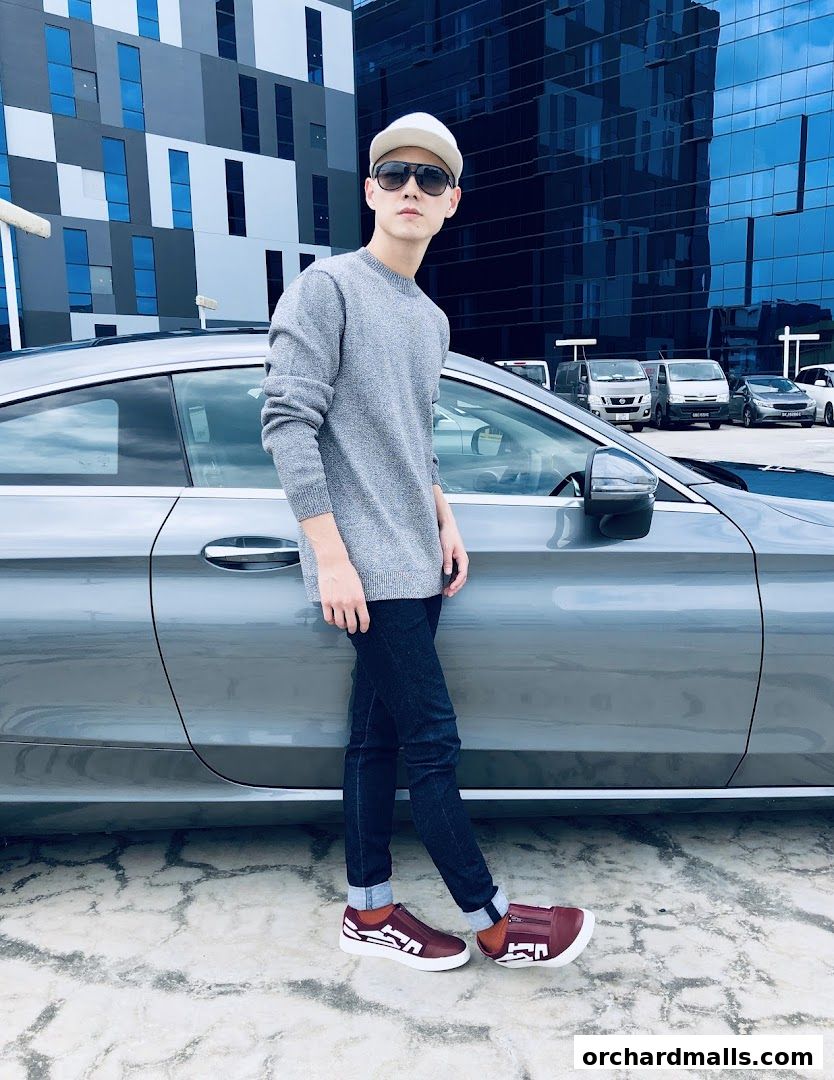 A man in a grey sweater and jeans poses next to a car with a modern building in the background.