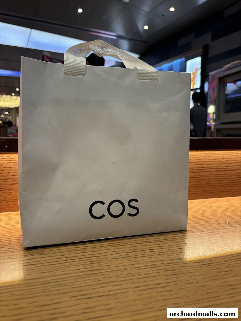 A white shopping bag with the COS logo sits on a wooden surface.