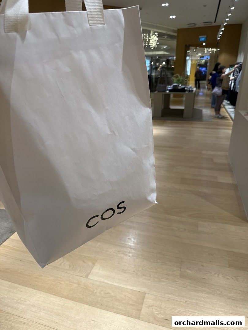 A white shopping bag with the COS logo in front of a blurred store interior.