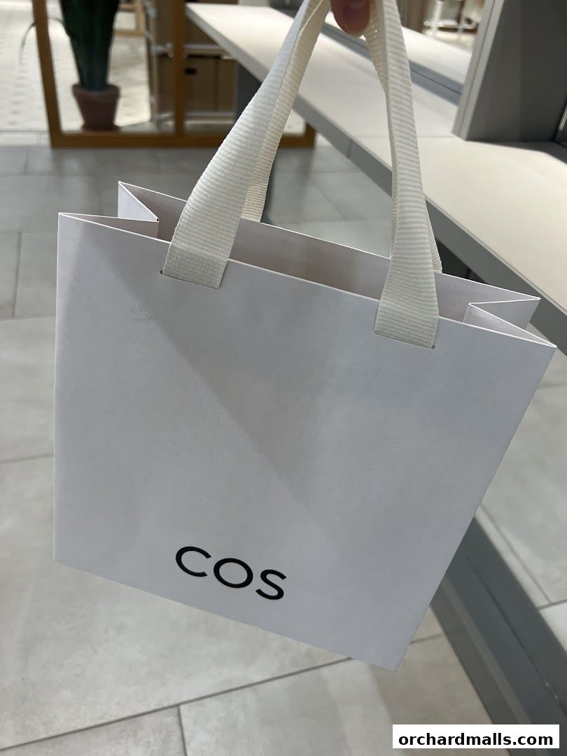 A white shopping bag with the COS logo is held up in front of a store interior.