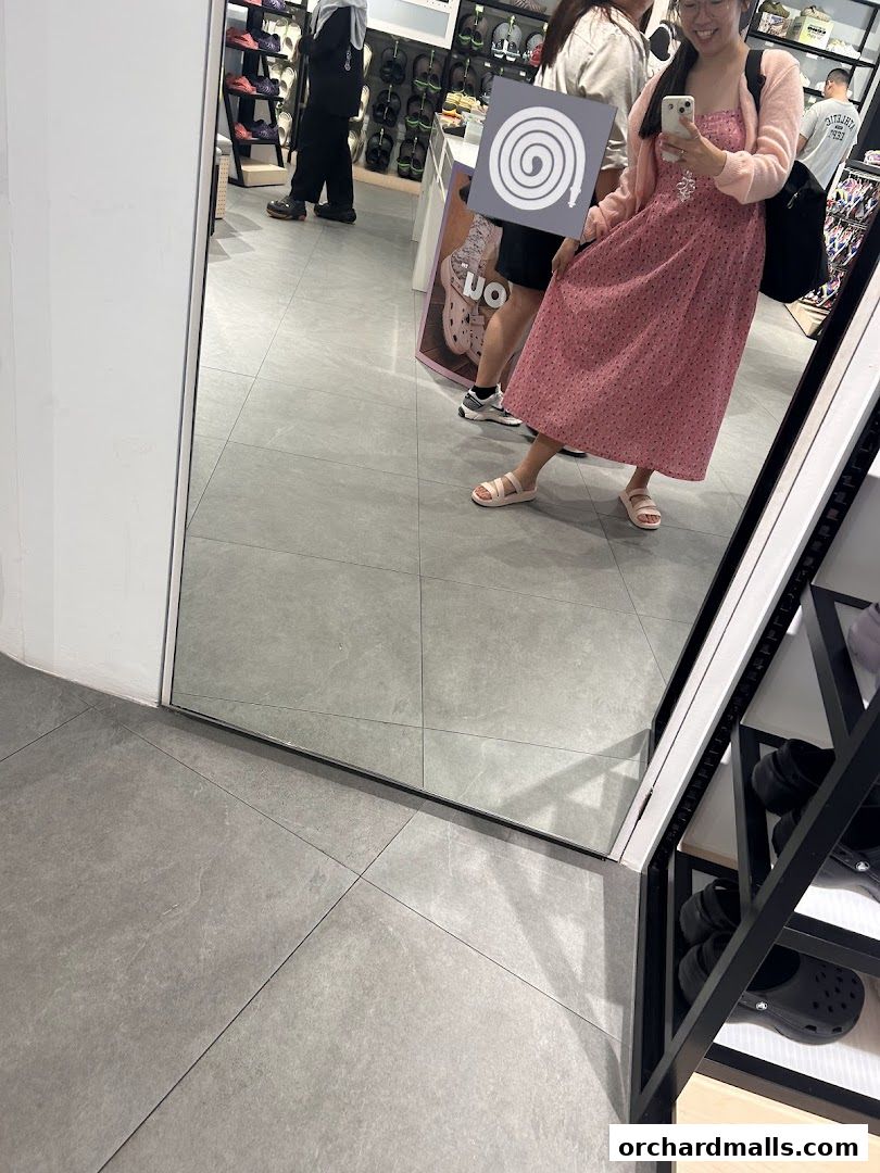 A woman in a pink dress poses in a Crocs store, reflecting in a large mirror.