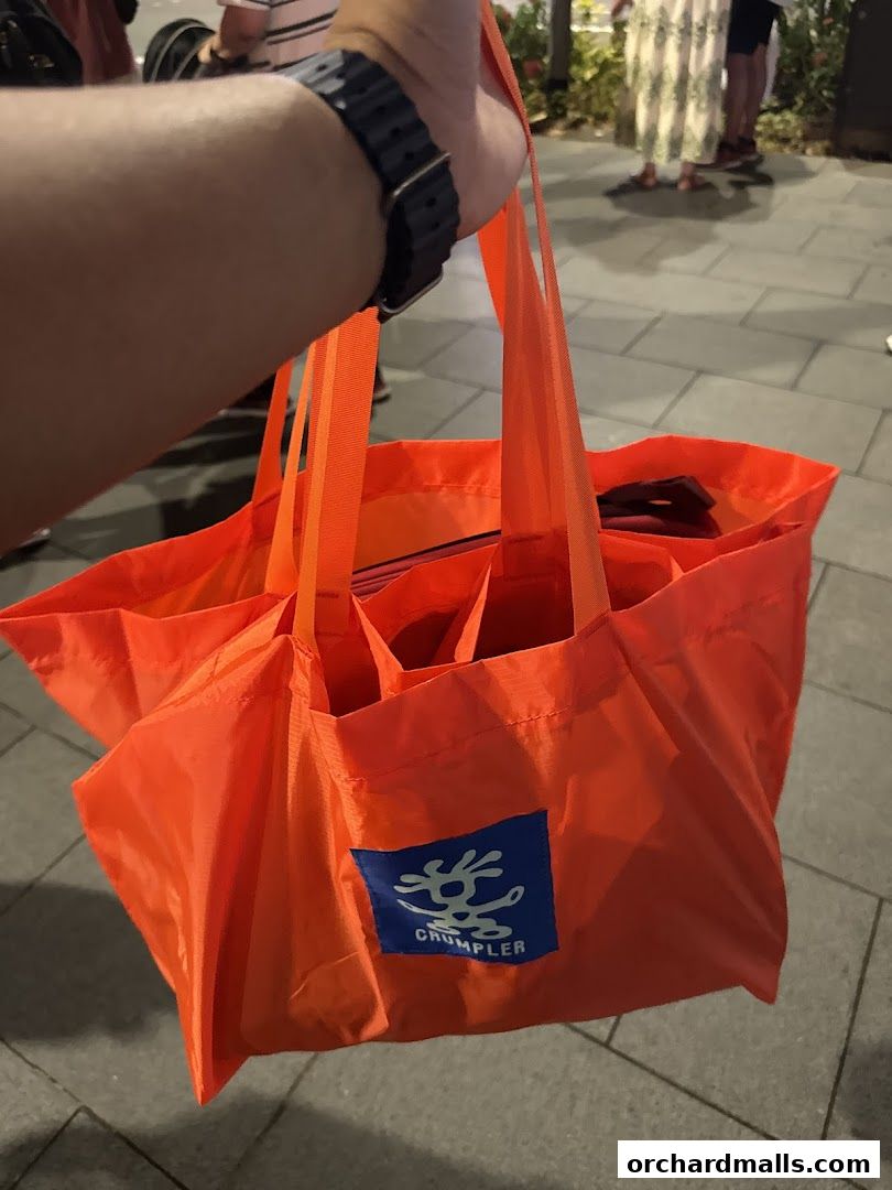 A hand holds a bright orange Crumpler tote bag with multiple compartments.