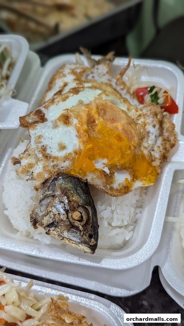 A close-up of a takeaway meal with rice, fried eggs, and a fried fish head.