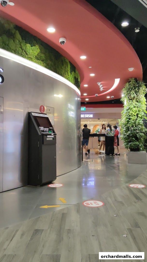 Interior view of a mall with an ATM, greenery, and a sushi restaurant entrance.