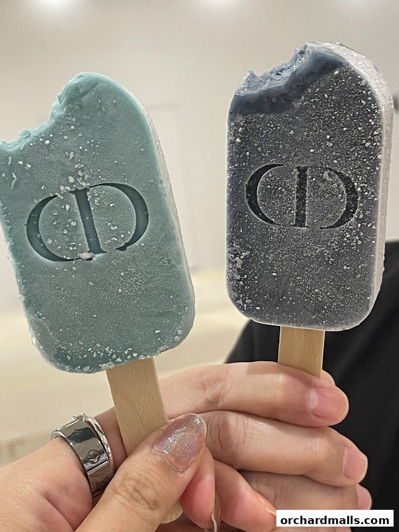 Two Dior-branded popsicles, one blue and one grey, are held by hands.