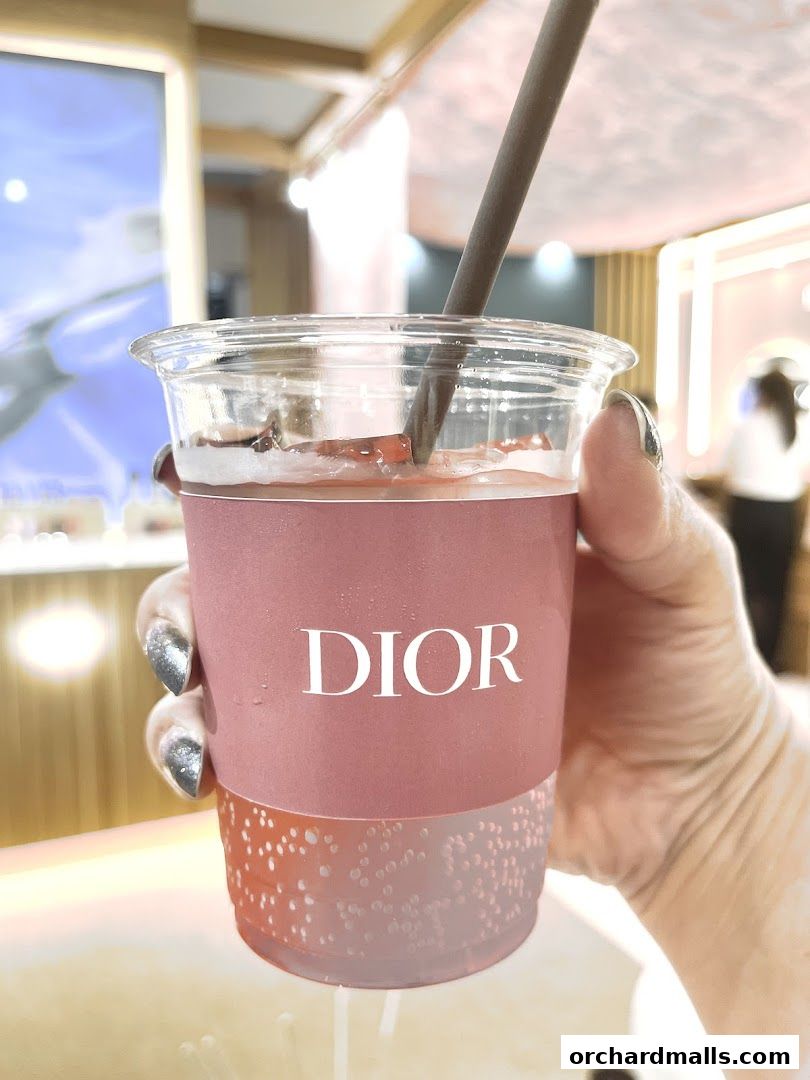 A hand holds a pink cup with the Dior logo, containing a beverage with a straw.