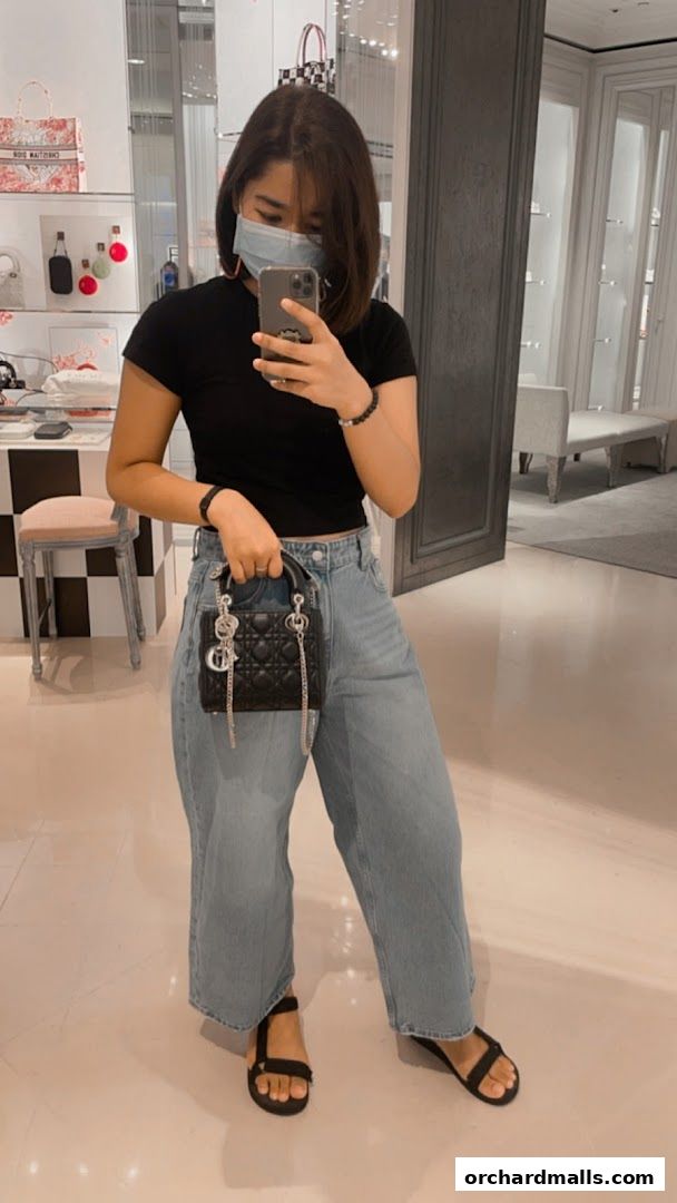 A woman in a black top and jeans poses with a Dior handbag inside a boutique.