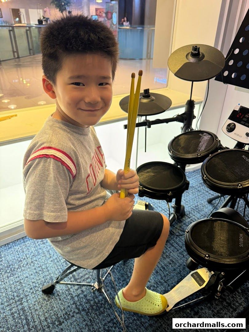 A young boy smiles while holding drumsticks next to a drum set.