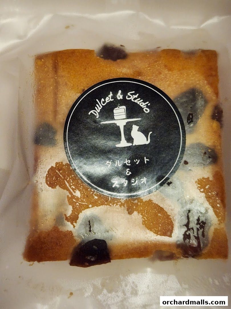 A slice of cake with dried fruit, packaged in plastic, with a "Dulcet & Studio" logo.