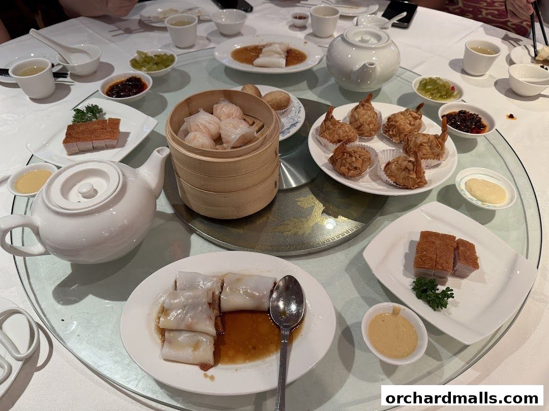 A table laden with various Chinese dishes, including dim sum, roasted meats, and rice noodle rolls.
