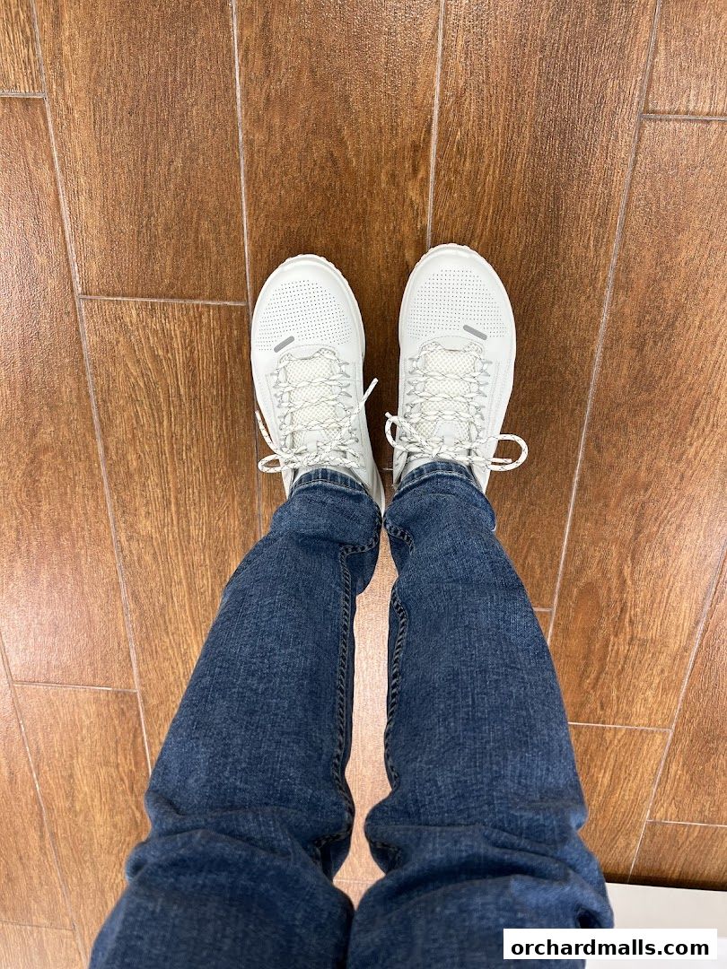 A person wearing white ECCO sneakers and blue jeans standing on a wooden floor.