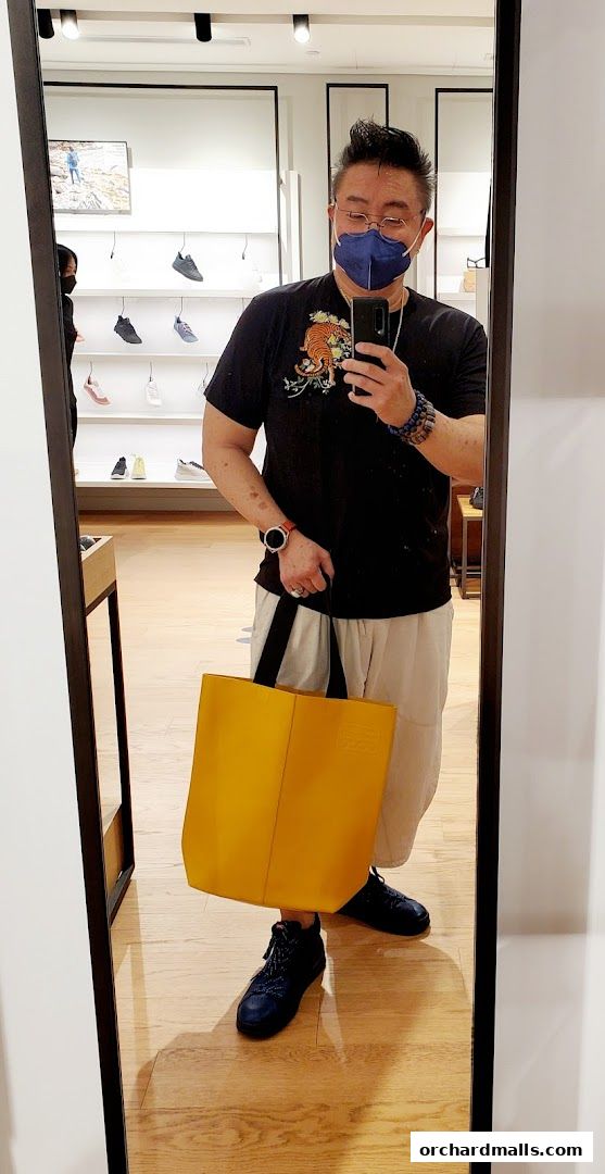 A man in a mask takes a mirror selfie holding a yellow tote bag in a shoe store.