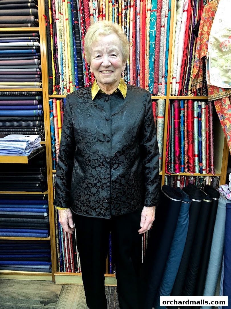 A woman stands in a tailor shop surrounded by shelves of colorful fabrics and rolls of material.