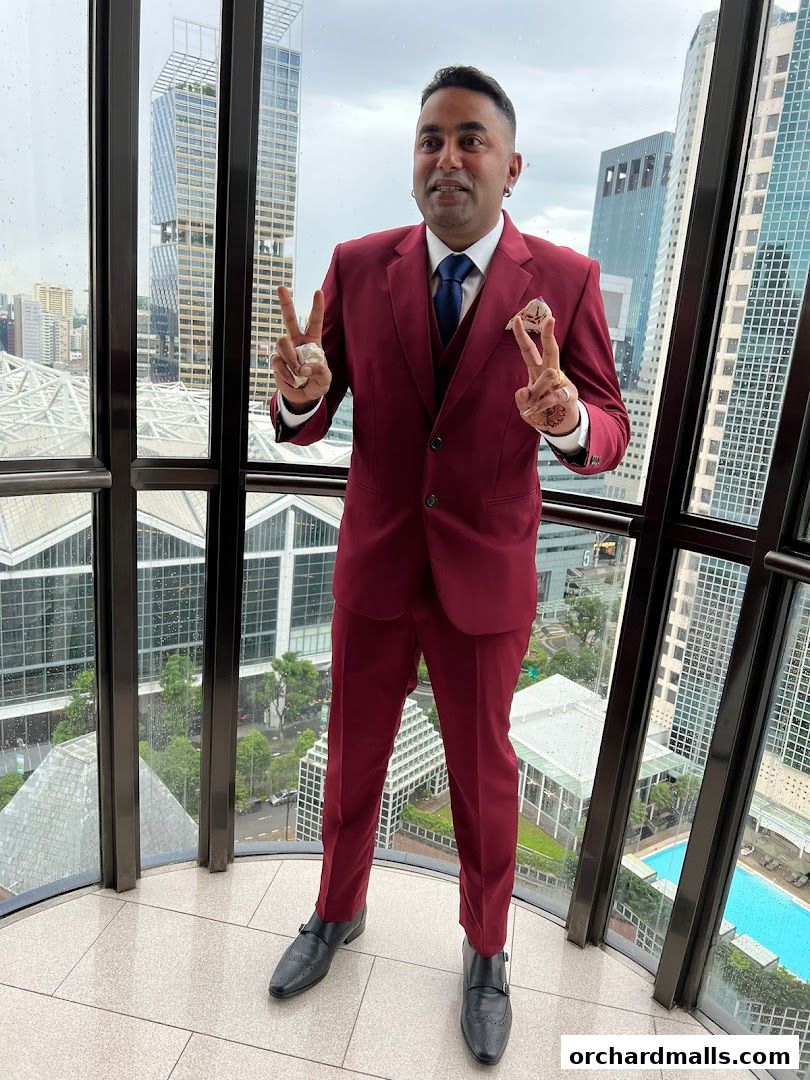 A man in a maroon suit stands by a window with a city skyline view.