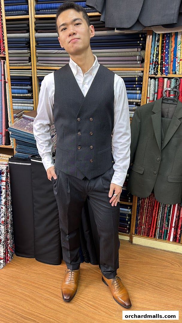 A man models a tailored suit in a fabric shop with shelves of colorful textiles.