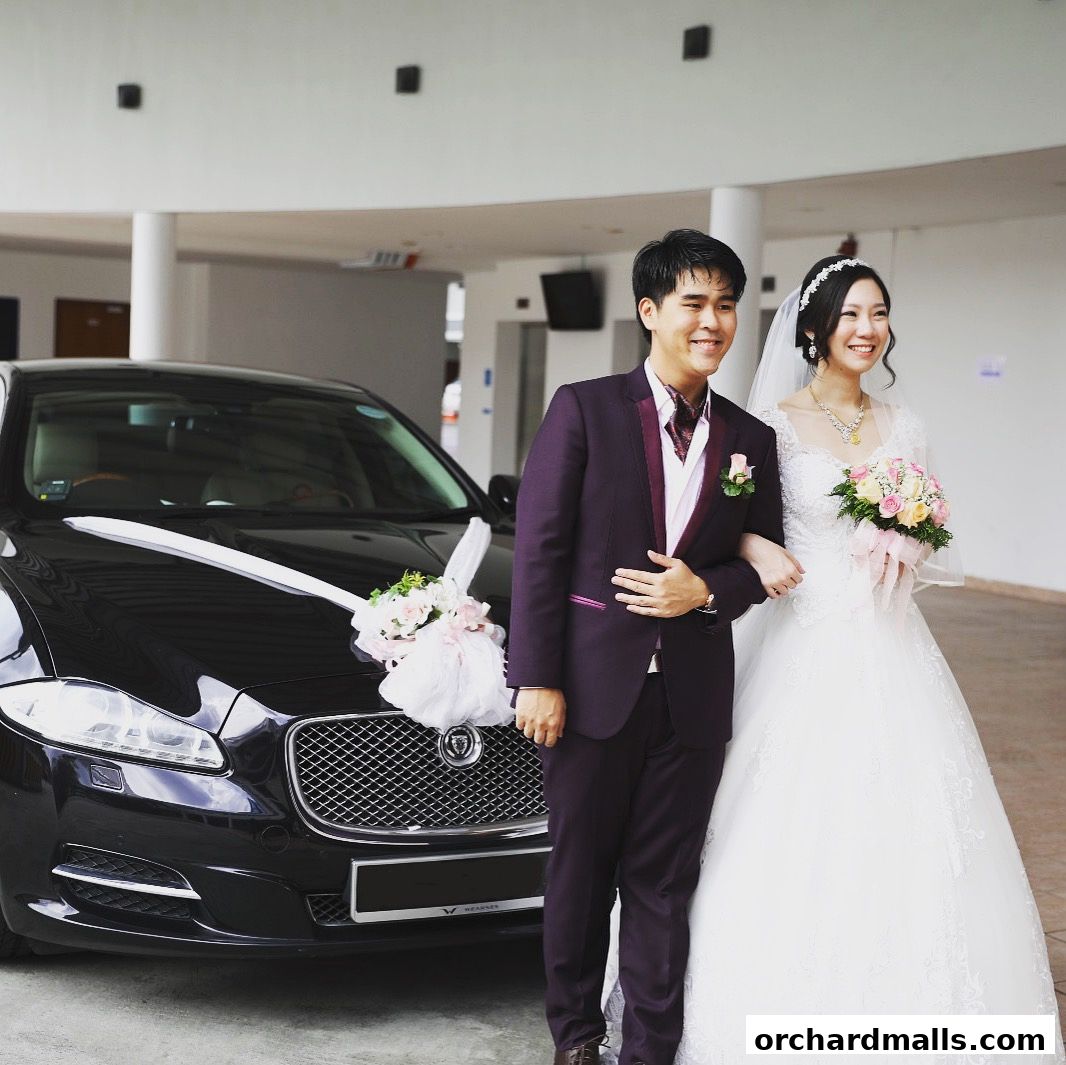 A smiling bride and groom pose next to a decorated black car on their wedding day.