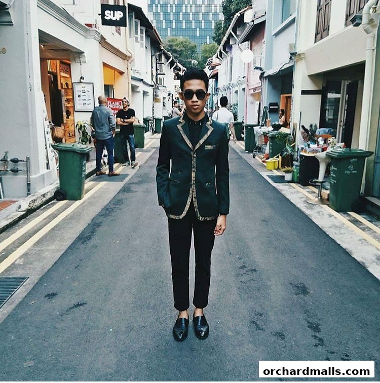 A man in a stylish suit stands in a narrow street lined with shops.