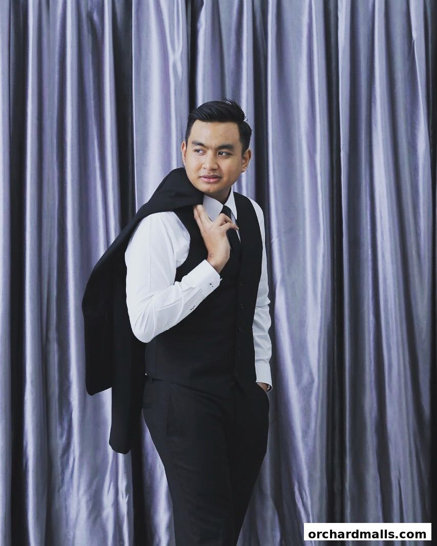 A man in a formal suit stands against a shimmering curtain backdrop.