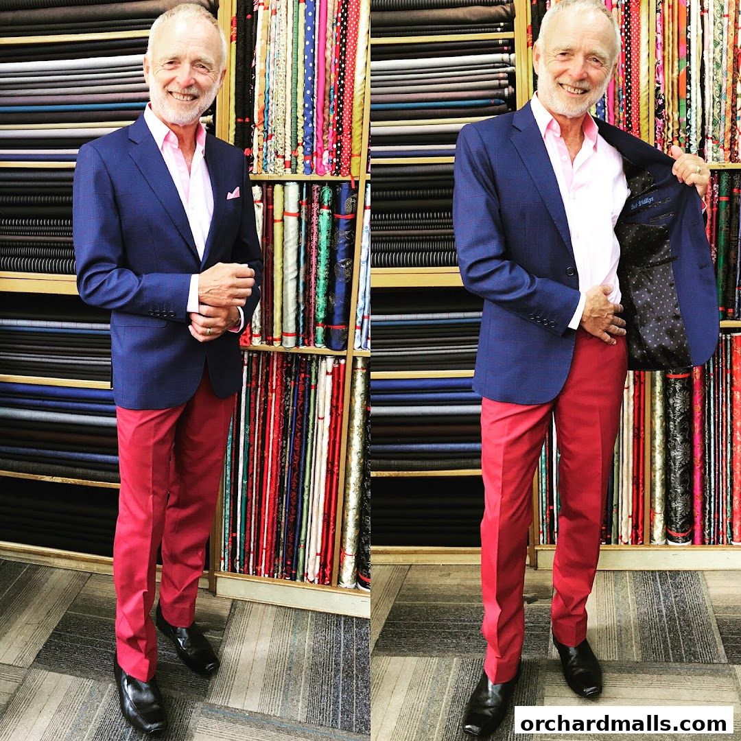 A man in a blue suit jacket and red pants stands in front of shelves of colorful fabrics.