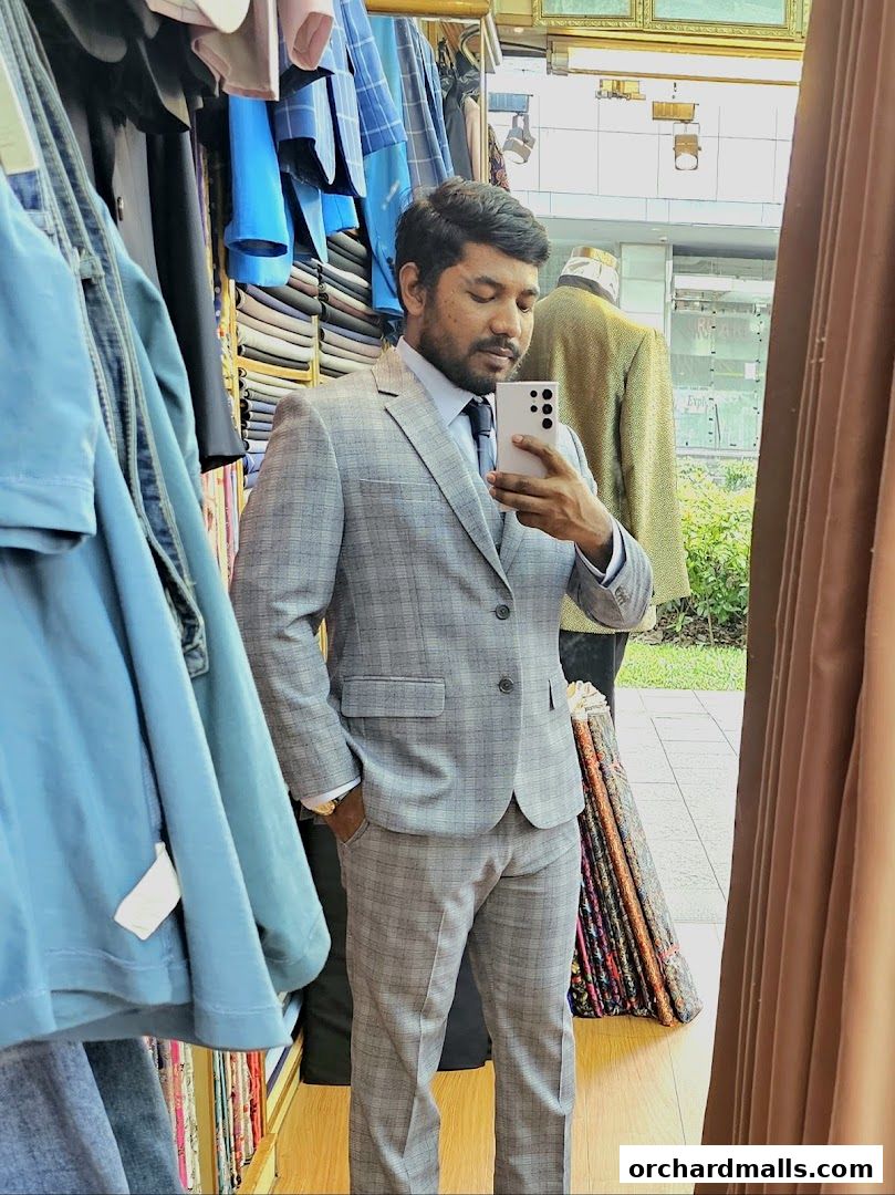 A man in a tailor shop tries on a grey plaid suit, surrounded by fabric rolls.