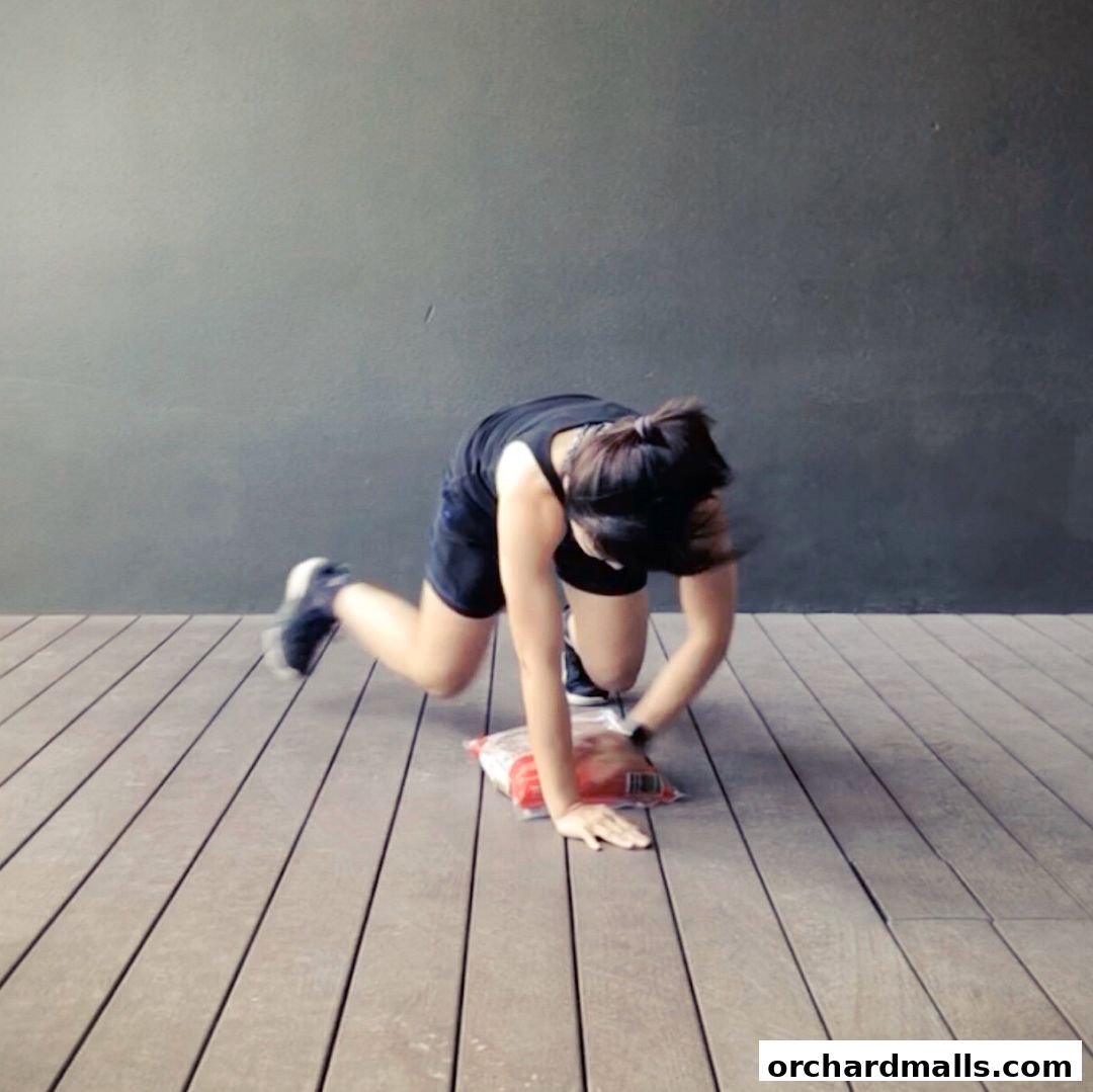 A woman in athletic wear performs an exercise on a wooden deck with a gray wall behind her.
