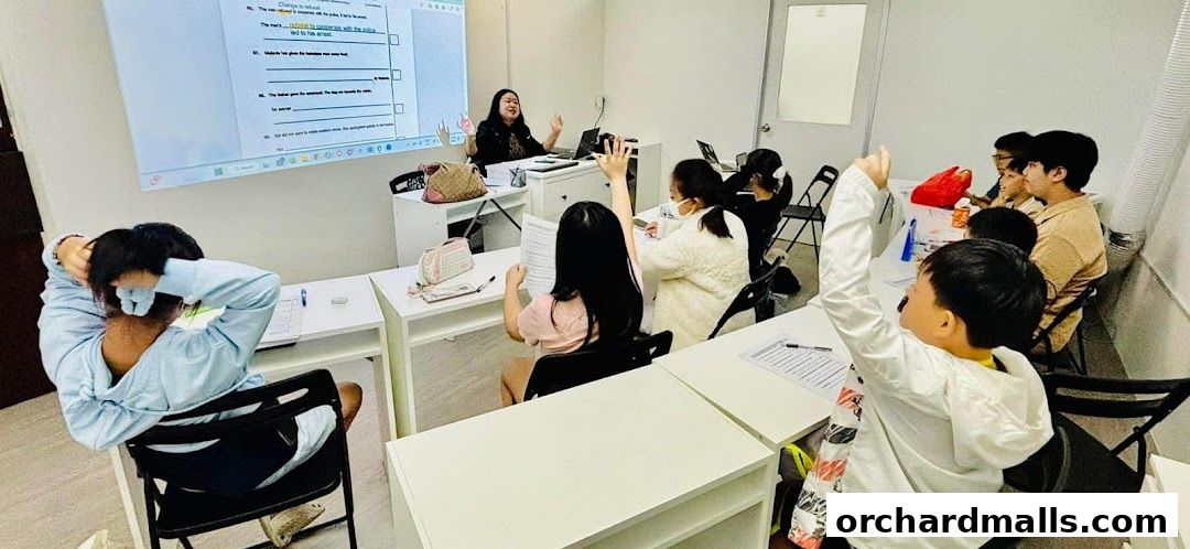 A teacher instructs students in a classroom setting, with many students raising their hands.
