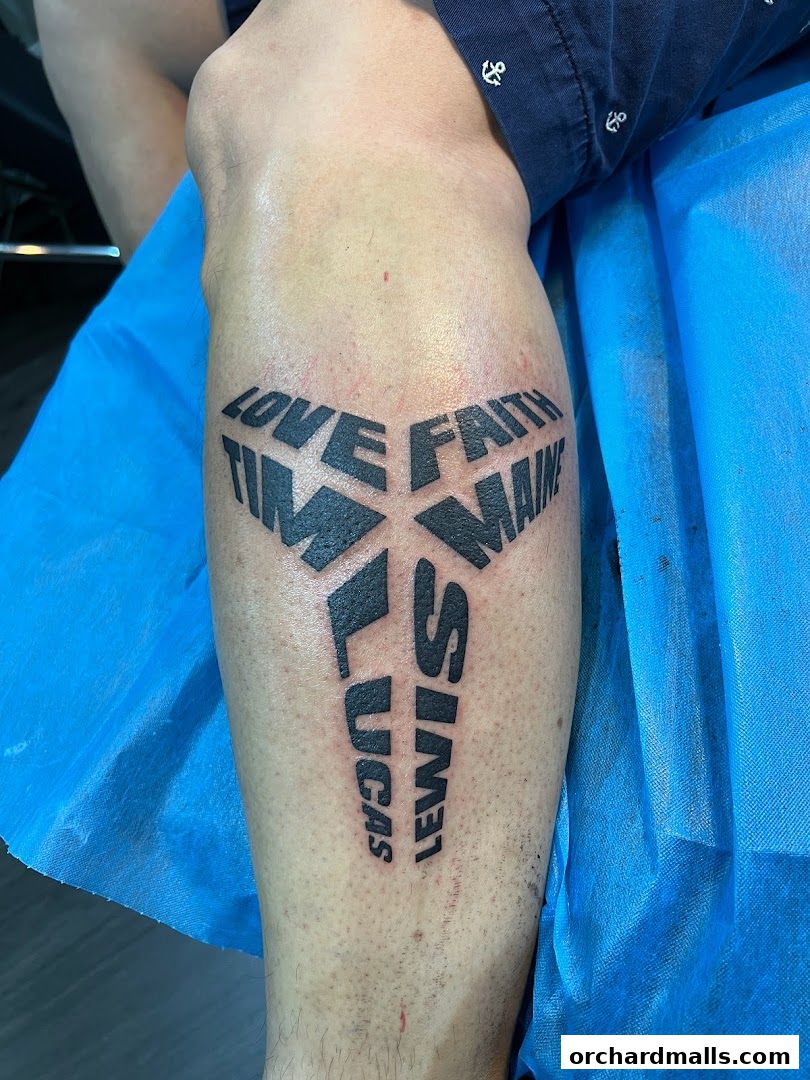 A close-up of a leg with a black tattoo featuring the words 'LOVE FAITH TIME MAINE LUCAS LEWIS'.