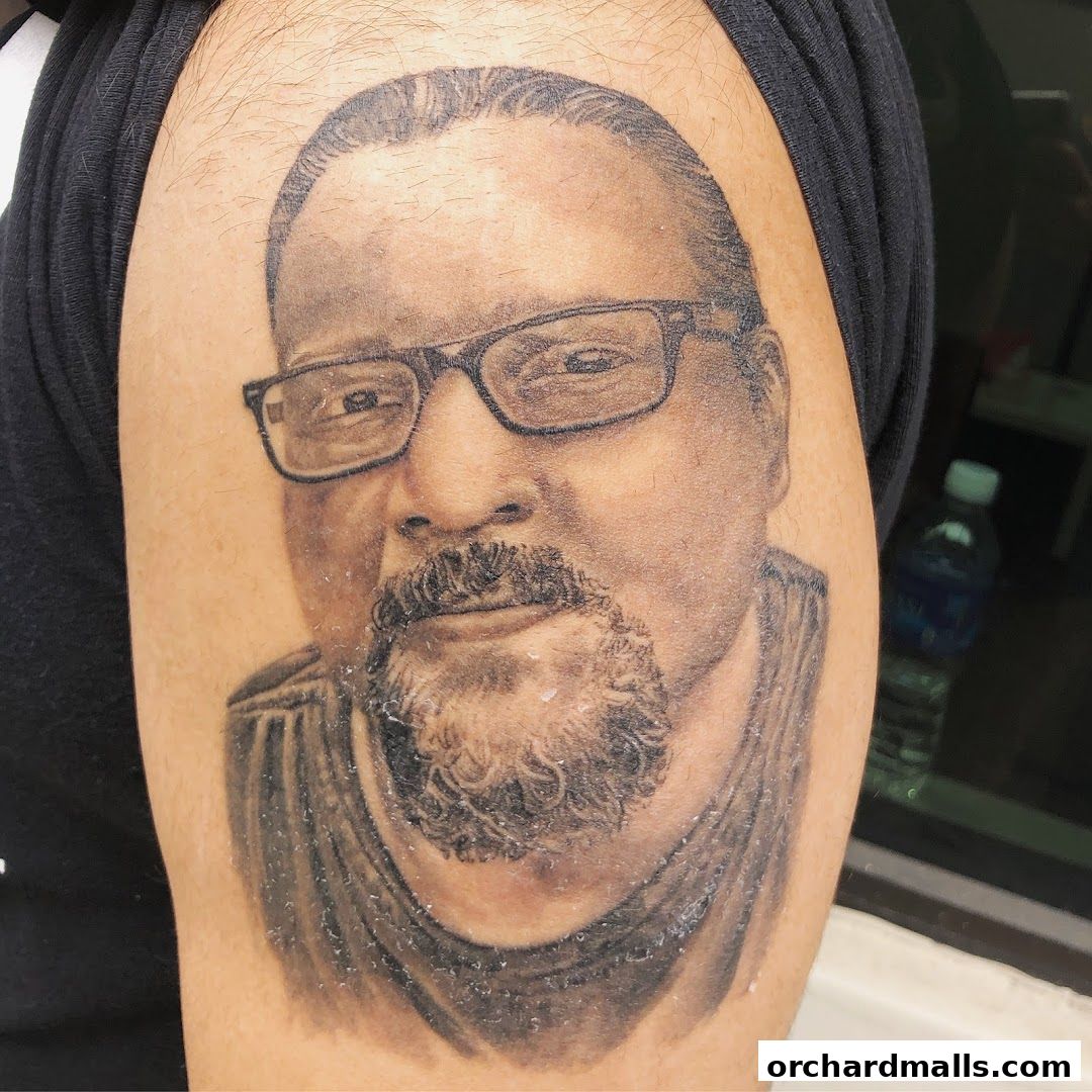 A detailed black and grey portrait tattoo of a man with glasses and a beard on an arm.