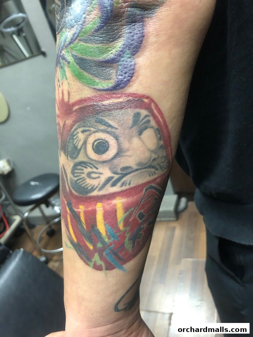 Close-up of a colorful Daruma doll tattoo on an arm, with other tattoo elements visible.