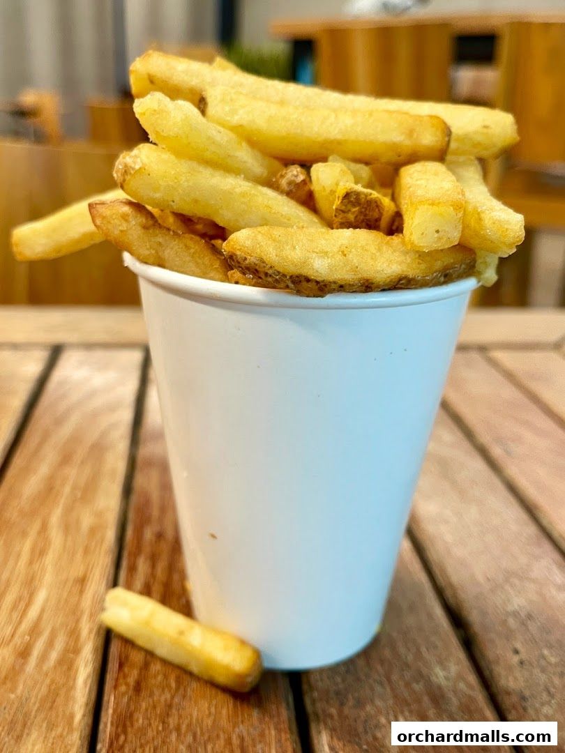 A white cup overflowing with golden, crispy French fries sits on a wooden table.