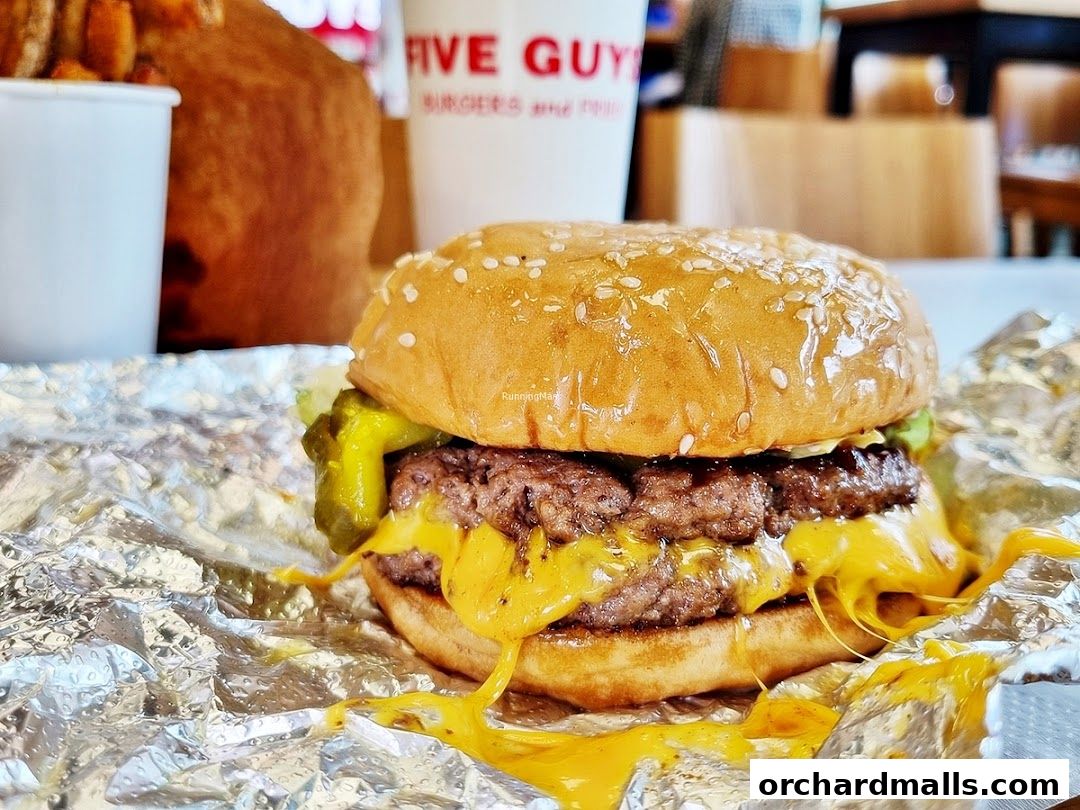 A juicy cheeseburger with melted cheese and pickles on foil, with fries and a drink in the background.