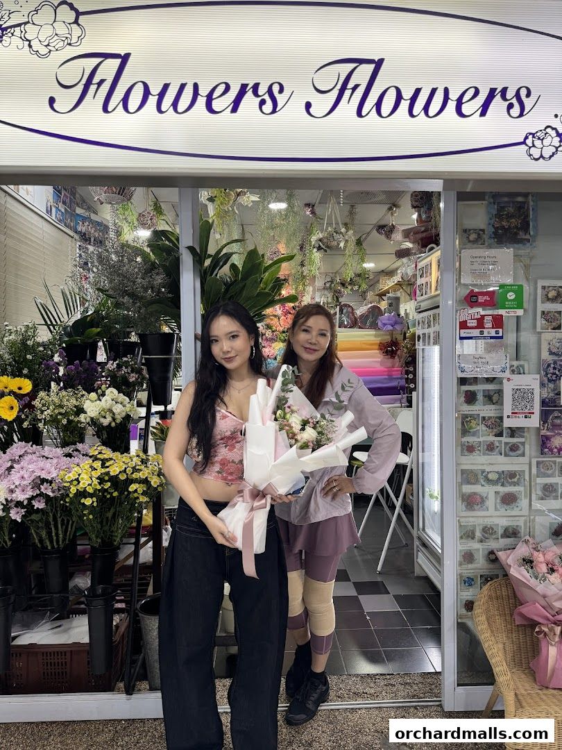 Two women stand outside a flower shop, holding bouquets.