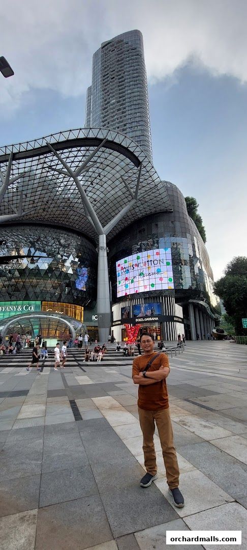 A man stands in front of a modern building with shops like Tiffany & Co. and Louis Vuitton.