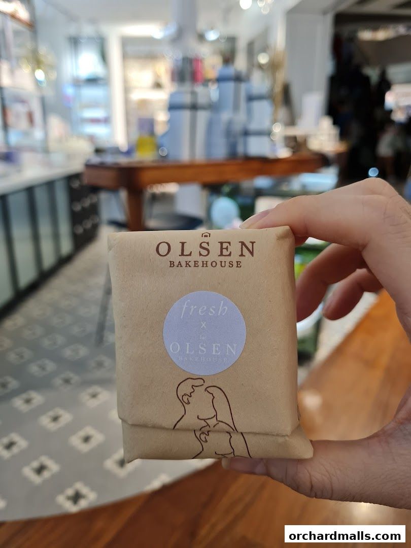 A hand holds a brown paper-wrapped item with 'Olsen Bakehouse' branding and a couple illustration.