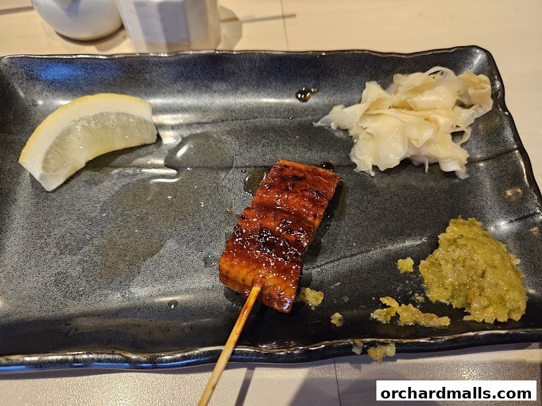 A skewer of glazed yakitori with lemon, pickled ginger, and wasabi on a dark plate.